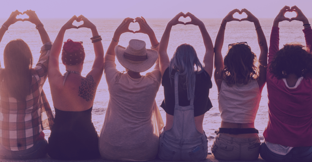 the backs of several women sitting in a line on the beach with their arms outstretched over their heads making hearts with their hands